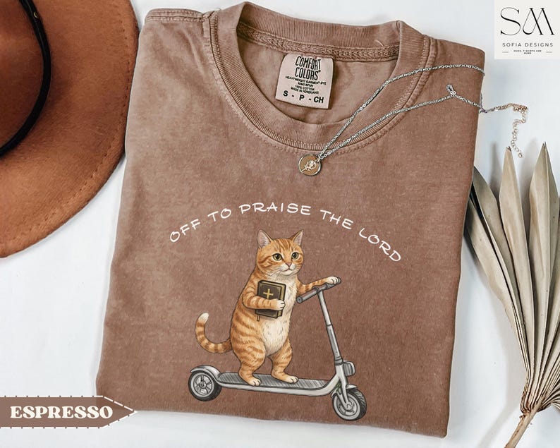 Puede incluir: Camiseta color espresso con el texto "Off to Praise the Lord". La camiseta presenta un gato de dibujos animados montado en un scooter y sosteniendo un libro. La camiseta tiene cuello redondo y mangas cortas. La etiqueta de la marca es visible.