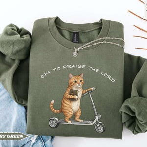 Puede incluir: Sudadera verde militar con el texto "OFF TO PRAISE THE LORD". El gr&aacute;fico presenta un gato atigrado sosteniendo un libro y montando un scooter. Un collar plateado est&aacute; drapeado sobre la sudadera. La sudadera est&aacute; doblada sobre unos vaqueros.