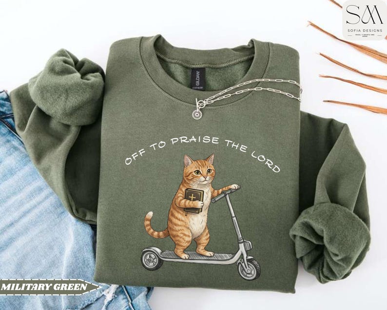 Puede incluir: Sudadera verde militar con el texto "OFF TO PRAISE THE LORD". El gr&aacute;fico presenta un gato atigrado sosteniendo un libro y montando un scooter. Un collar plateado est&aacute; drapeado sobre la sudadera. La sudadera est&aacute; doblada sobre unos vaqueros.