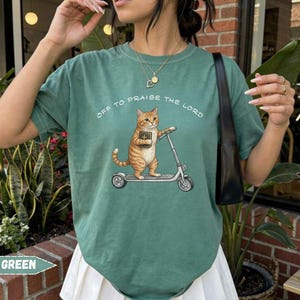 Christian cat graphic t-shirt featuring orange tabby on scooter with Bible, funny faith based cat lover tee, aesthetic Comfort Colors shirt gift