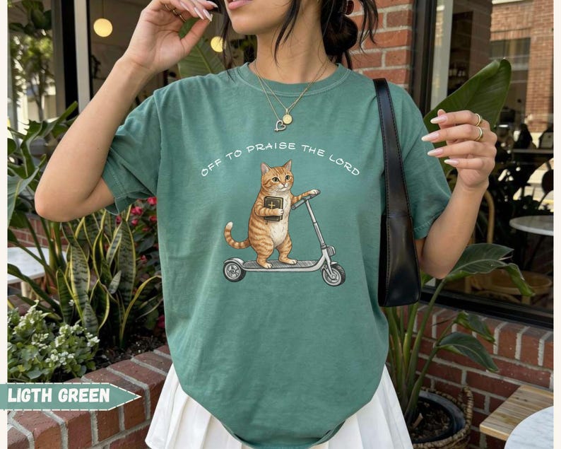 Christian cat graphic t-shirt featuring orange tabby on scooter with Bible, funny faith based cat lover tee, aesthetic Comfort Colors shirt gift