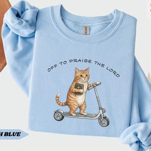 Puede incluir: Sudadera azul claro con la frase "OFF TO PRAISE THE LORD". El dise&ntilde;o presenta un gato atigrado naranja con una biblia en un patinete. La sudadera tiene cuello redondo y mangas largas.