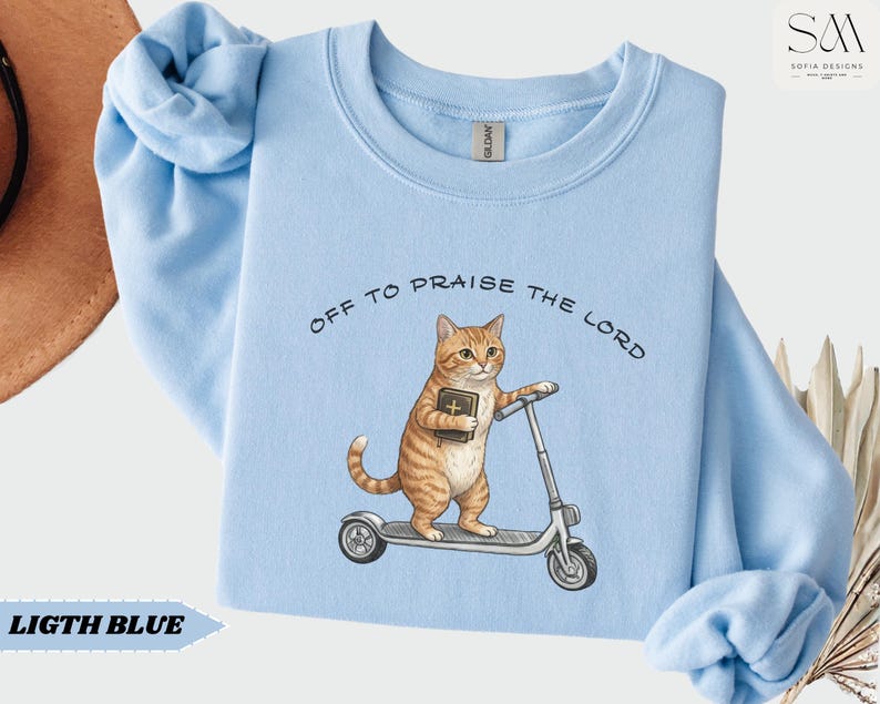 Puede incluir: Sudadera azul claro con la frase "OFF TO PRAISE THE LORD". El dise&ntilde;o presenta un gato atigrado naranja con una biblia en un patinete. La sudadera tiene cuello redondo y mangas largas.