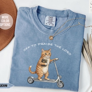 Puede incluir: Camiseta azul vaquero con el texto "OFF TO PRAISE THE LORD". La camiseta presenta un gr&aacute;fico de un gato atigrado naranja sosteniendo un libro y montando un scooter. Un collar dorado est&aacute; drapeado sobre la camiseta.