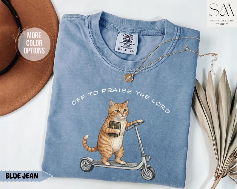 Puede incluir: Camiseta azul vaquero con el texto "OFF TO PRAISE THE LORD". La camiseta presenta un gr&aacute;fico de un gato atigrado naranja sosteniendo un libro y montando un scooter. Un collar dorado est&aacute; drapeado sobre la camiseta.