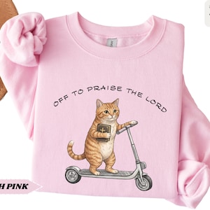 Puede incluir: Sudadera rosa claro con el texto "OFF TO PRAISE THE LORD". El dise&ntilde;o presenta un gato atigrado naranja montado en un patinete y sosteniendo un libro. La sudadera es de color rosa claro.