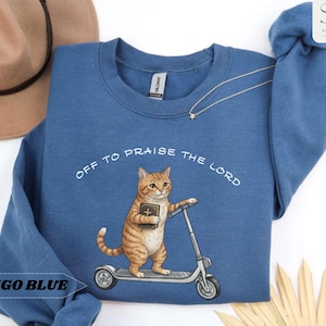 Puede incluir: Sudadera azul &iacute;ndigo con el texto "OFF TO PRAISE THE LORD". El dise&ntilde;o presenta un gato atigrado montado en un patinete con un libro. La sudadera es de cuello redondo.