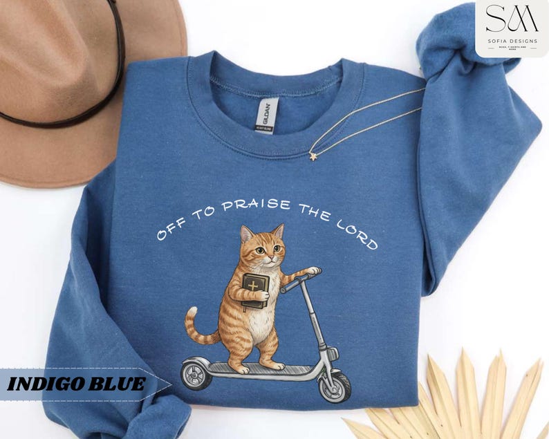 Puede incluir: Sudadera azul &iacute;ndigo con el texto "OFF TO PRAISE THE LORD". El dise&ntilde;o presenta un gato atigrado montado en un patinete con un libro. La sudadera es de cuello redondo.