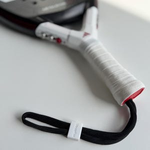 Può includere: Una racchetta da padel bianca e nera con un'impugnatura bianca e un cinturino da polso nero. L'impugnatura è avvolta con nastro bianco e il cinturino ha una chiusura bianca con la scritta "StrapFix". La racchetta ha un accento rosso.