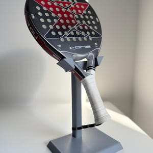 Peut inclure: Une raquette de padel NOX X-ONE noire et rouge avec une poignée blanche est présentée sur un support gris. La raquette a une surface perforée et un motif en forme de X rouge. Le support est à base carrée et a un support vertical.