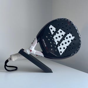 Puede incluir: Una raqueta de pádel en blanco y negro con el texto "METALBONE" y el logotipo de Adidas. La raqueta se muestra sobre un soporte negro. El mango es blanco con una correa de muñeca negra.