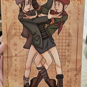 May include: A rectangular art print featuring two figures in a dance pose, wearing berets, scarves, and dark clothing with brown boots. The background is tan with the text "The Changeling" at the top and "Clara Saburoba" vertically on both sides.