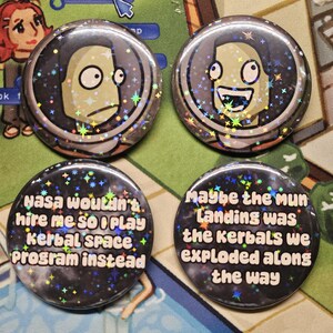 May include: Four circular pin-back buttons featuring holographic star patterns. Two buttons show cartoon characters in space helmets with wide eyes. The other two buttons feature the text "NASA wouldn't hire me so I play Kerbal Space Program instead" and "maybe the mun landing was the kerbals we exploded along the way."