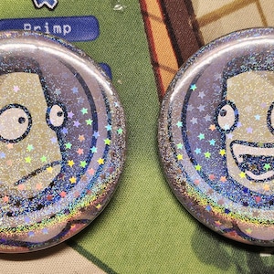 May include: Two circular pin-back buttons featuring a holographic star-patterned finish. Each button displays a cartoon character with a pale face and large eyes inside a dark blue circular frame. One character has a neutral expression, while the other has a wide, open-mouthed smile.