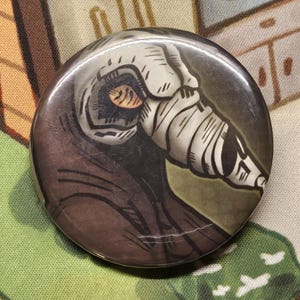 May include: A circular pin-back button featuring a stylized illustration of a figure wearing a white, cone-shaped mask with dark, hatched line details. The figure is dressed in a dark brown garment, and the design uses bold black outlines.