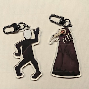 May include: Two acrylic keychains with black metal clasps. One keychain features a black figure with a white mask, the other a plague doctor with a long beak and dark cloak. Both keychains are on a white surface.