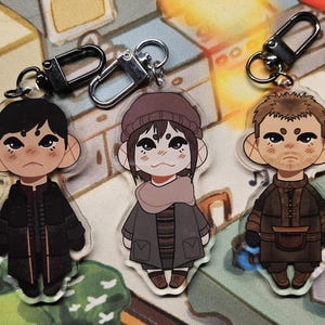 May include: Three acrylic keychains featuring cartoon-style characters with large ears and expressive faces. Each charm includes a silver-toned metal lobster clasp. The characters wear dark, layered winter clothing, including coats, a beanie, and a scarf.