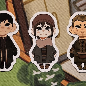 May include: Three die-cut stickers featuring stylized cartoon characters with large ears. The left character wears a black coat, the center character wears a brown beanie and scarf with a dark coat, and the right character wears a brown jacket.