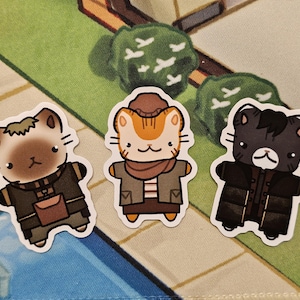 May include: Three die-cut stickers featuring cartoon cats dressed in layered outfits. The left cat is tan and brown with a satchel, the center cat is orange and white with a brown hat and scarf, and the right cat is black and grey with a dark coat.