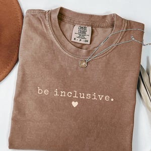 Peut inclure: Un t-shirt marron avec les mots "be inclusive." et un cœur. Le t-shirt a un col rond et des manches courtes. Un collier argenté avec un pendentif est drapé sur le t-shirt. Fabriqué par Comfort Colors.