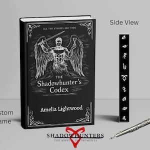 May include: A black book titled "The Shadowhunter's Codex" with a white illustration of a winged figure holding a sword. A bookmark with symbols and a silver pen are next to the book. The book cover also has the name "Amelia Lightwood".