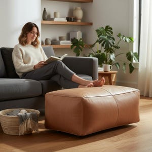 Op de afbeelding: Een rechthoekige, lichtbruine lederen voetenbank met een centrale naad, geplaatst voor een grijze bank. De voetenbank heeft een glad, gestructureerd oppervlak en staat op een houten vloer, wat een stevige steun biedt voor de voeten.