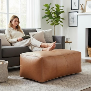 Puede incluir: Un otomán rectangular de cuero color canela con costuras visibles, que presenta una superficie lisa y de color sólido con un diseño de perfil bajo. El otomán está colocado frente a un sofá gris, ofreciendo una superficie estable.