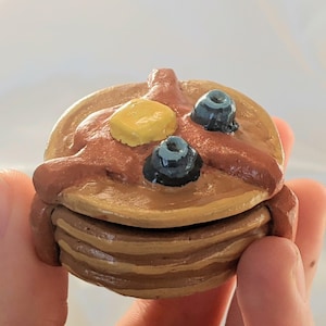 May include: A miniature stack of pancakes with butter and blueberries. The pancakes are a light brown colour, with a darker brown syrup, a yellow pat of butter, and two dark blue blueberries on top. The pancakes are held in a person's hand.