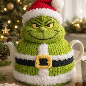 Puede incluir: Una caprichosa funda de tetera verde tejida en forma de personaje con un gorro de Papá Noel rojo. La funda tiene un ribete blanco, un cinturón negro y una hebilla amarilla. La tetera está sobre una mesa de madera con galletas.