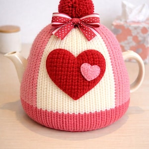 Könnte beinhalten: Ein gestrickter Teewärmer in Rosa, Weiß und Rot mit einem Herzmotiv. Der Wärmer hat einen roten Bommel und eine rot-weiß gepunktete Schleife. Ein kleines rosa Herz liegt auf einem größeren roten Herz.