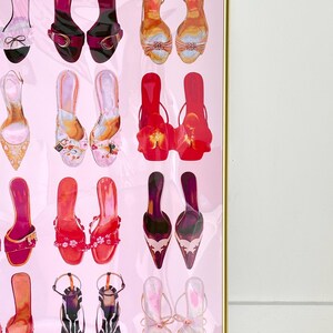 May include: A framed wall art print featuring a grid of various high-heeled sandals and shoes in shades of pink, red, orange, and black. The collection includes strappy heels, buckled designs, and floral embellished footwear on a light pink background.