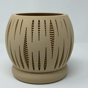 Könnte beinhalten: Beigefarbener, kugelförmiger Pflanztopf mit dekorativem Ausschnittmuster. Der Pflanztopf steht auf einem abgerundeten Sockel. Das Ausschnittmuster erzeugt einen geschichteten Effekt und verleiht dem Stück visuelles Interesse. Geeignet für den Innen- und Außenbereich.