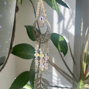 Puede incluir: Un adorno colgante decorativo con forma de media luna de metal dorado, un amuleto de mano de Hamsa y hebras de cuentas colgantes. El adorno incluye cuentas transparentes e iridiscentes y un dije en forma de hoja en la parte inferior.