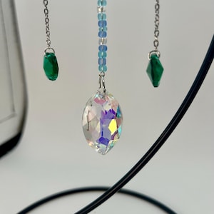 Puede incluir: Un cristal colgante decorativo con refracción de luz iridiscente. Está suspendido de una cadena plateada y adornado con cuentas azules y transparentes. Dos colgantes de cristal verde adicionales cuelgan de cadenas plateadas.