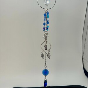 Peut inclure: Un attrape-soleil décoratif avec un cristal transparent et des perles bleues. L'attrape-soleil a un cadre et des accents en métal argenté, dont un petit charme de hibou et des pendentifs en forme de feuilles. Les perles varient en taille et en nuance de bleu.
