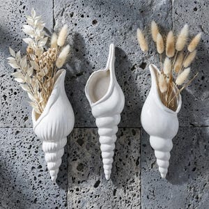 May include: Three white ceramic seashell-shaped wall vases. Each vase is filled with dried beige and white decorative plants. The vases are mounted on a textured gray wall, creating a coastal or nautical theme.