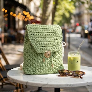May include: A pale green crocheted backpack with a flap and gold-tone clasp sits on a marble table. Tortoiseshell sunglasses and a glass of green drink with ice and a straw are also on the table.