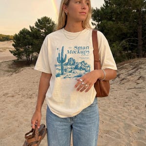 May include: Cream-colored t-shirt with a blue desert landscape graphic and the text "Smart Mockups Lab." The person is wearing light blue jeans and holding brown leather sandals. A brown leather purse is on the shoulder.
