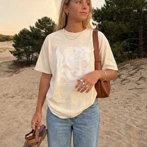 May include: Cream-colored t-shirt with a white floral design and the text "SMART MOCK". The person is wearing blue jeans and holding brown sandals. A brown leather bag is over the shoulder. A rainbow is visible in the background.