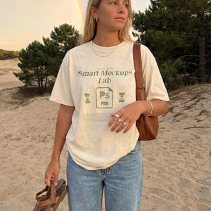 May include: Cream-colored t-shirt with the text "Smart Mockups Lab" in green. The shirt is paired with light blue jeans and brown leather sandals. A brown leather purse is worn over the shoulder. The background includes a rainbow and trees.