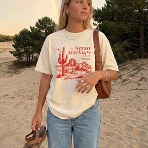 May include: Cream-colored t-shirt with a red desert landscape graphic and the text "Smart Mockups Lab." The shirt is paired with light blue jeans. A brown leather bag is worn over the shoulder, and brown sandals are held in the hand.