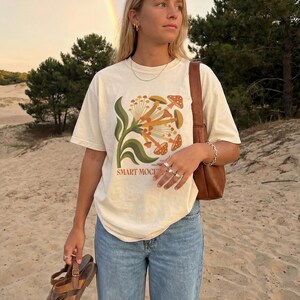 May include: Cream-colored t-shirt with a mushroom graphic and the text "SMART MOCI". The shirt is paired with blue jeans and brown sandals. A brown purse is worn over the shoulder. A rainbow is visible in the background.