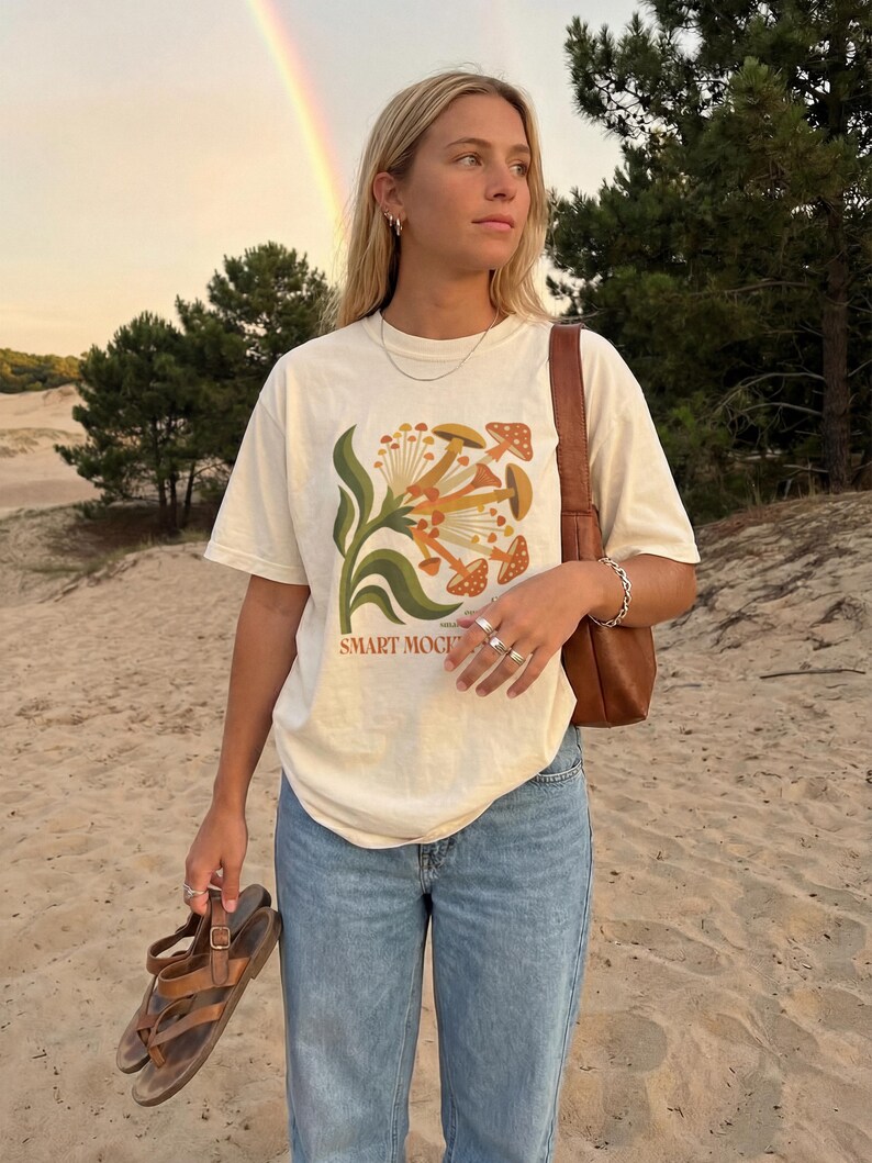 May include: Cream-colored t-shirt with a mushroom graphic and the text "SMART MOCI". The shirt is paired with blue jeans and brown sandals. A brown purse is worn over the shoulder. A rainbow is visible in the background.
