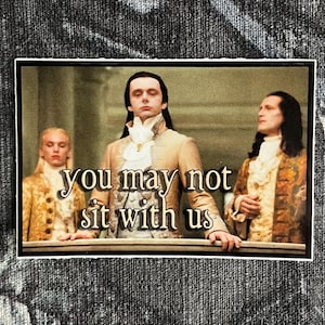 May include: A sticker featuring a scene with three people in historical attire. The text "you may not sit with us" is overlaid on the image. The sticker is set against a dark, patterned background.
