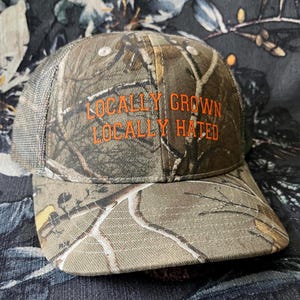 May include: A camouflage trucker hat with a mesh back and a curved brim. The hat features the orange text "LOCALLY GROWN LOCALLY HATED" across the front. The hat is designed with a woodland camouflage pattern.