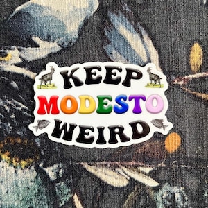 May include: A white sticker with the text "Keep Modesto Weird" in black. The word "Modesto" is in rainbow colors. There are two black and white goat illustrations and two black and white fish illustrations.
