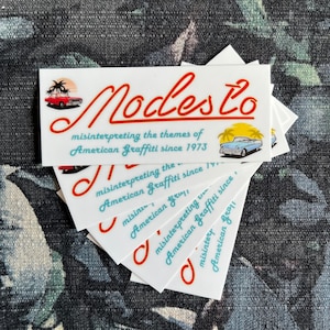 May include: A stack of white stickers with a red and blue logo that says "Modesto" and the text "misinterpreting the themes of American Graffiti since 1973". The logo features a red car and palm trees.