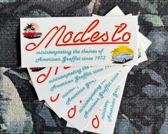 Op de afbeelding: Een stapel witte stickers met een rood en blauw logo dat "Modesto" zegt en de tekst "misinterpreting the themes of American Graffiti since 1973". Het logo toont een rode auto en palmbomen.