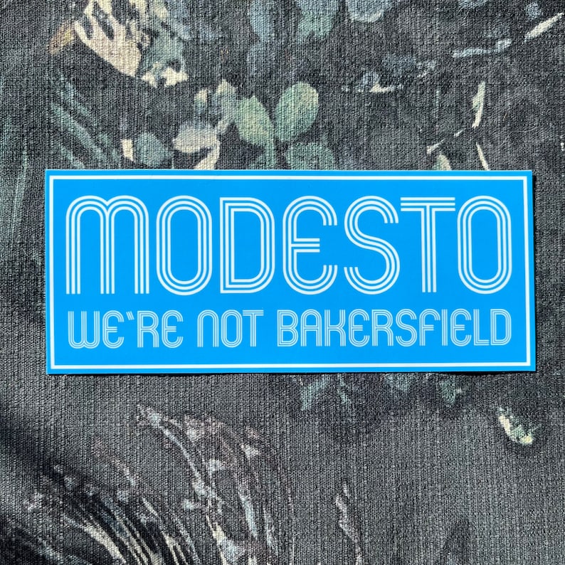 May include: A blue rectangular sticker with white text that reads "MODESTO WE'RE NOT BAKERSFIELD". The text is in a retro font style.