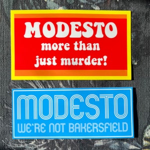 May include: Two stickers with the word "MODESTO" in a retro font. The top sticker is red with a yellow border and says "more than just murder!" The bottom sticker is blue with white text and says "WE'RE NOT BAKERSFIELD".